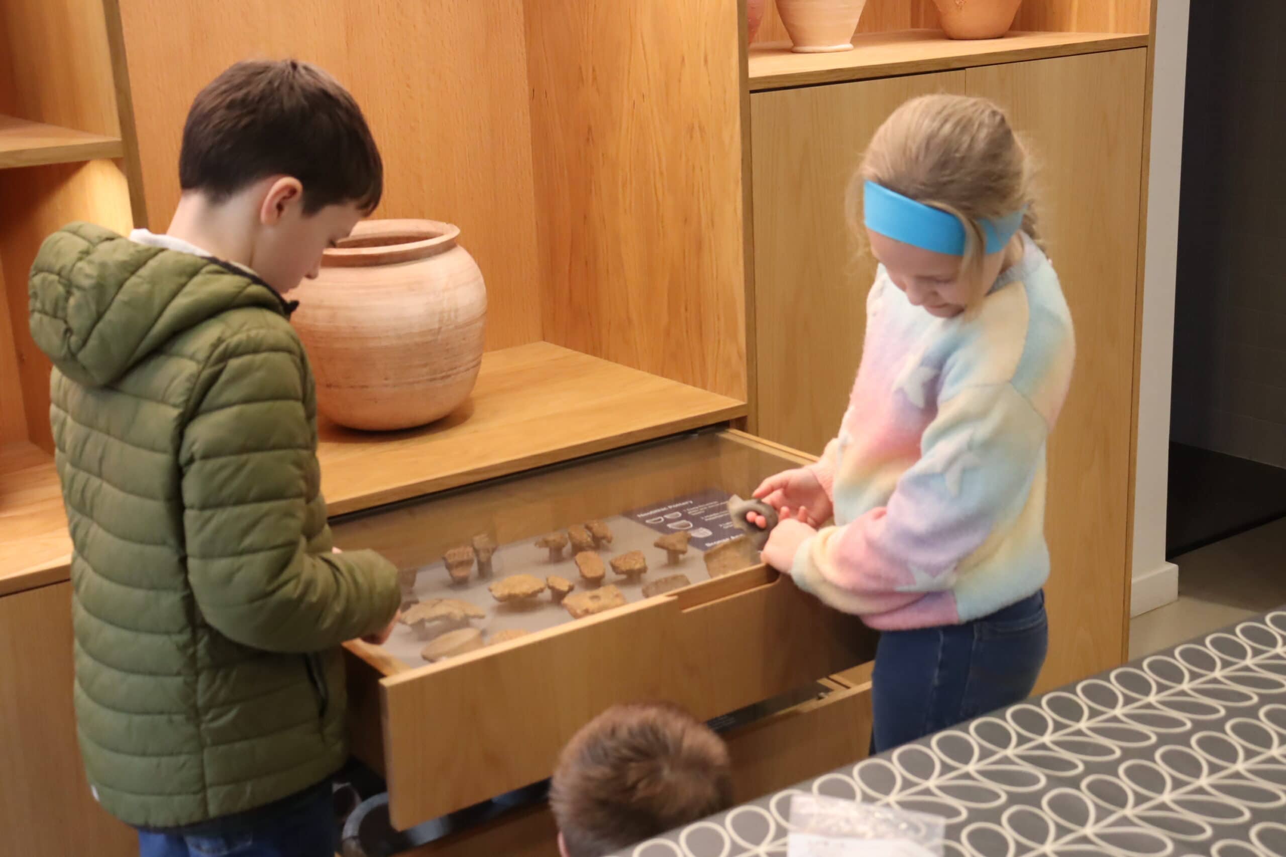 Two young people in a wood-paneled museum exhibition room look at a display case of pottery fragments.