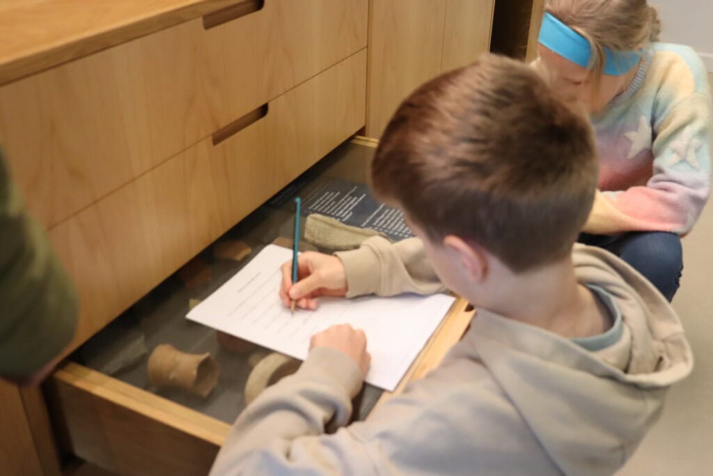A young person sketches archaeological objects in a draw with a glass cover