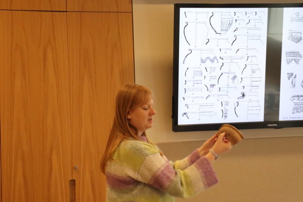A woman holds up a piece of archaeological pottery in front of a screen showing lots of illustrations of pottery fragments.