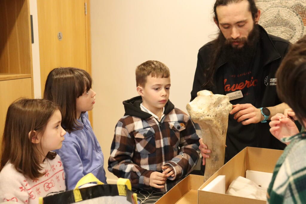 A group of young people and one adult looking at a large animal bone in a museum