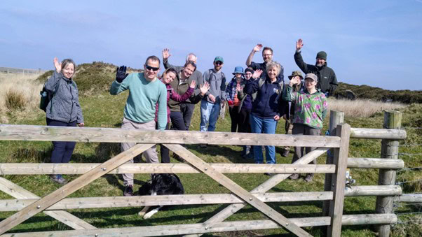 A group of adults and children waving from behind a wooden gate in a field on a sunny day.