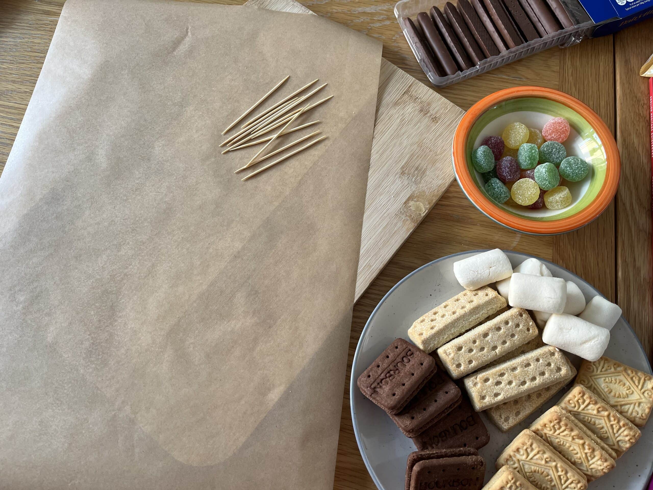 A sheet of greaseproof paper laid over a wooden chopping board, next to a plate of biscuits and a small bowl of colourful sweets