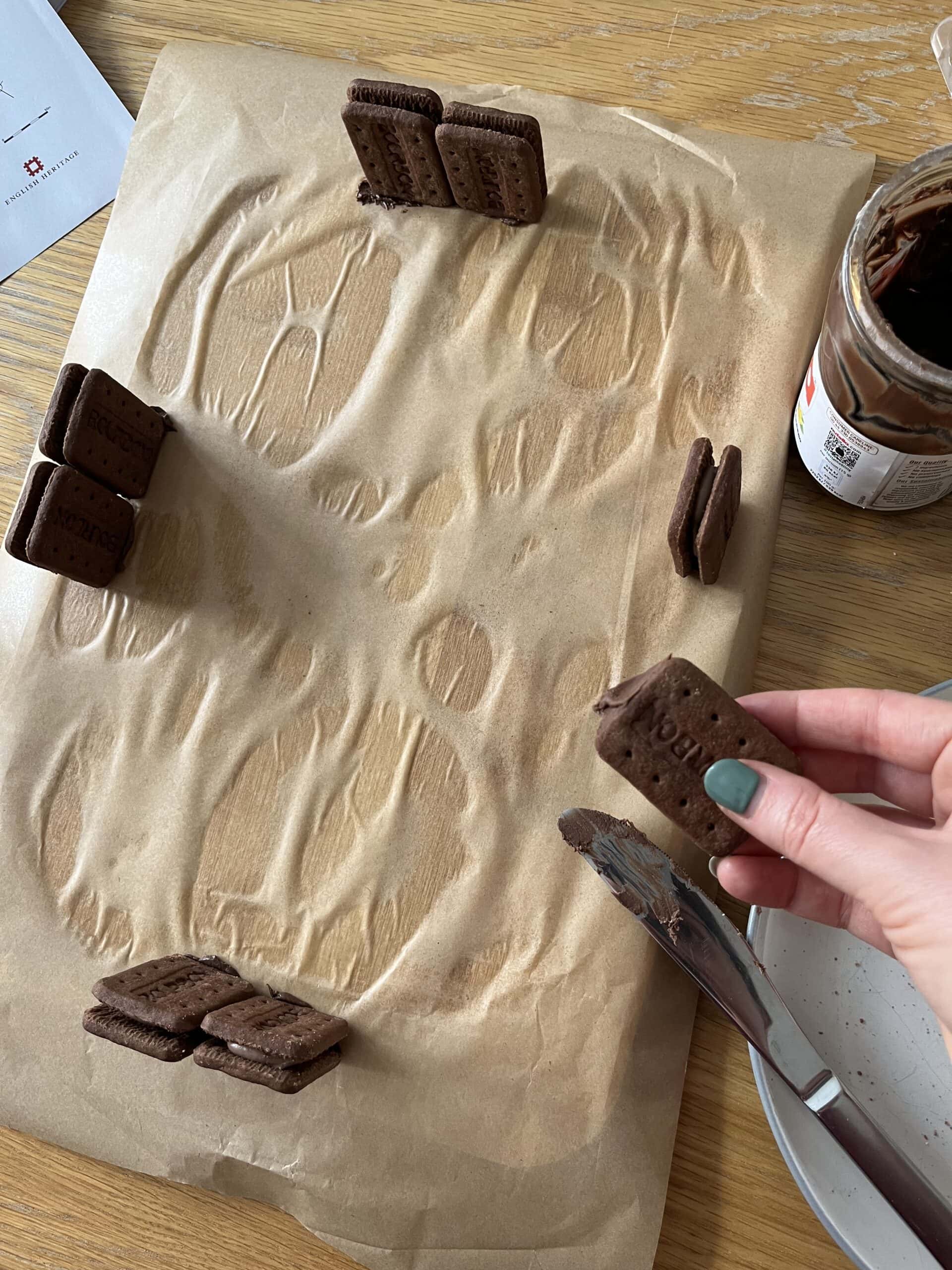 Four pairs of bourbon biscuits stuck upright onto a wooden chopping board covered with brown paper, next to a jar of chocolate spread.
