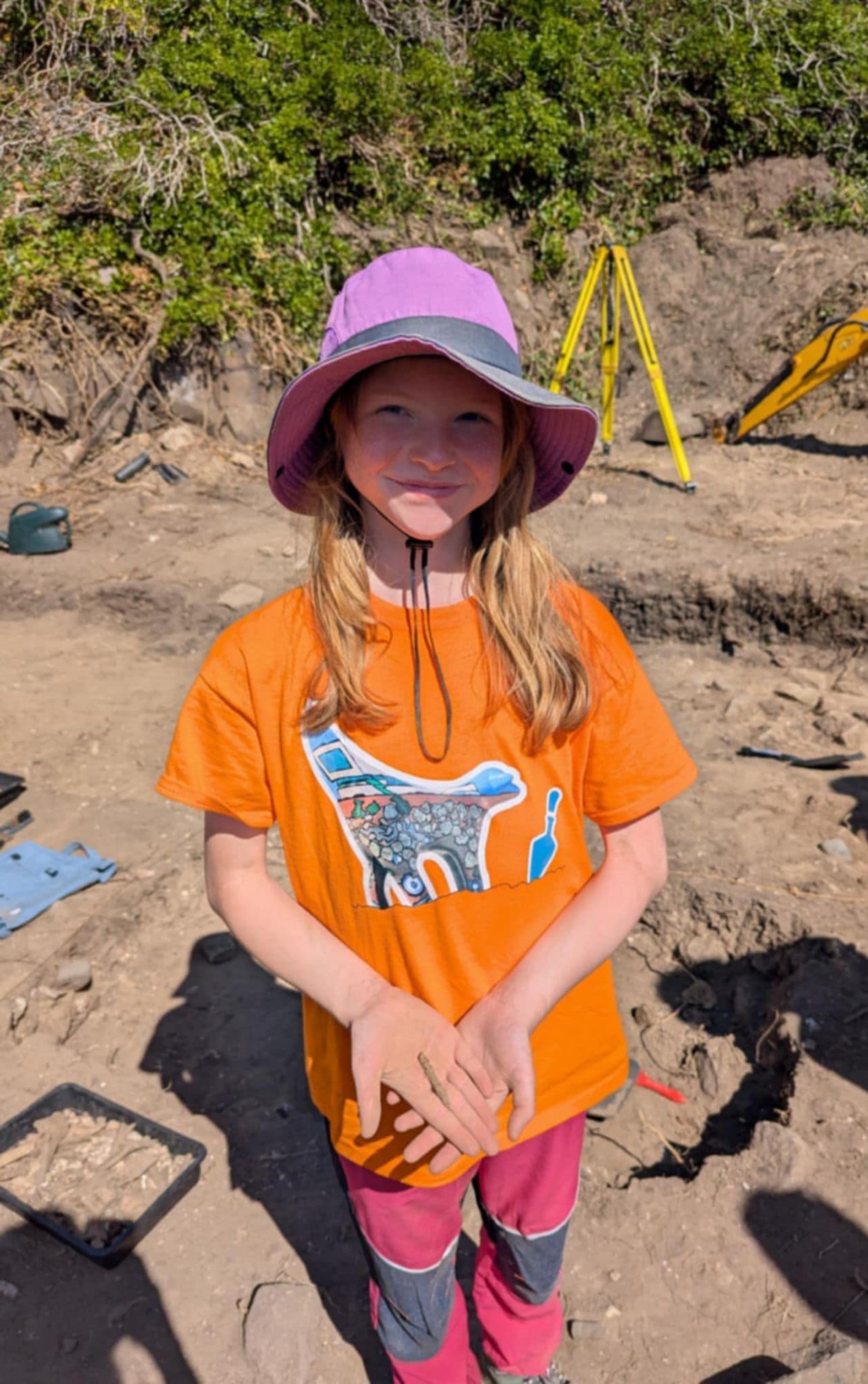 Child with long blonde hair wearng a pink bucet hat and orange t.shirt stood in trench