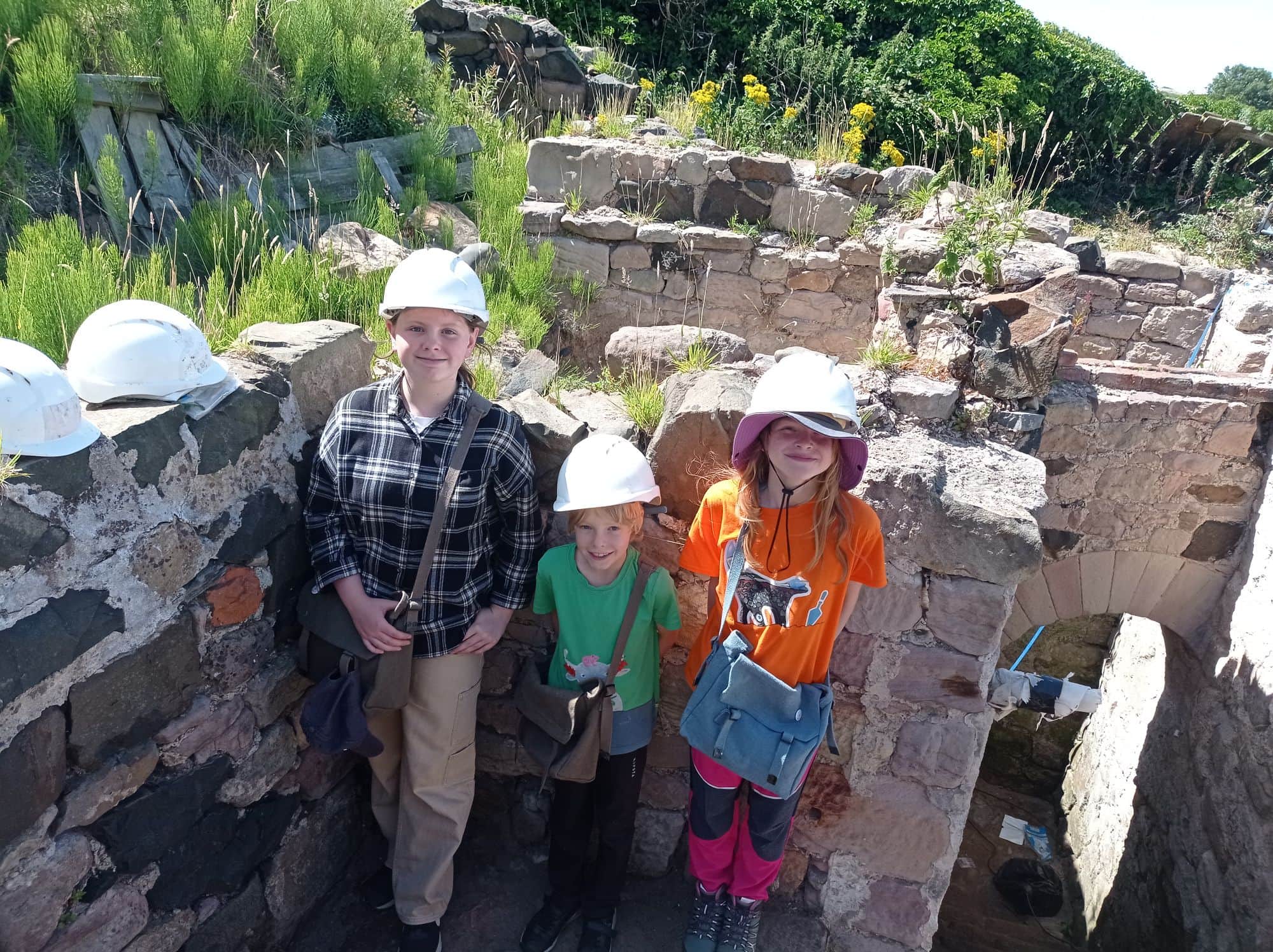 Three young people with white hard hats stood in front of stone builing