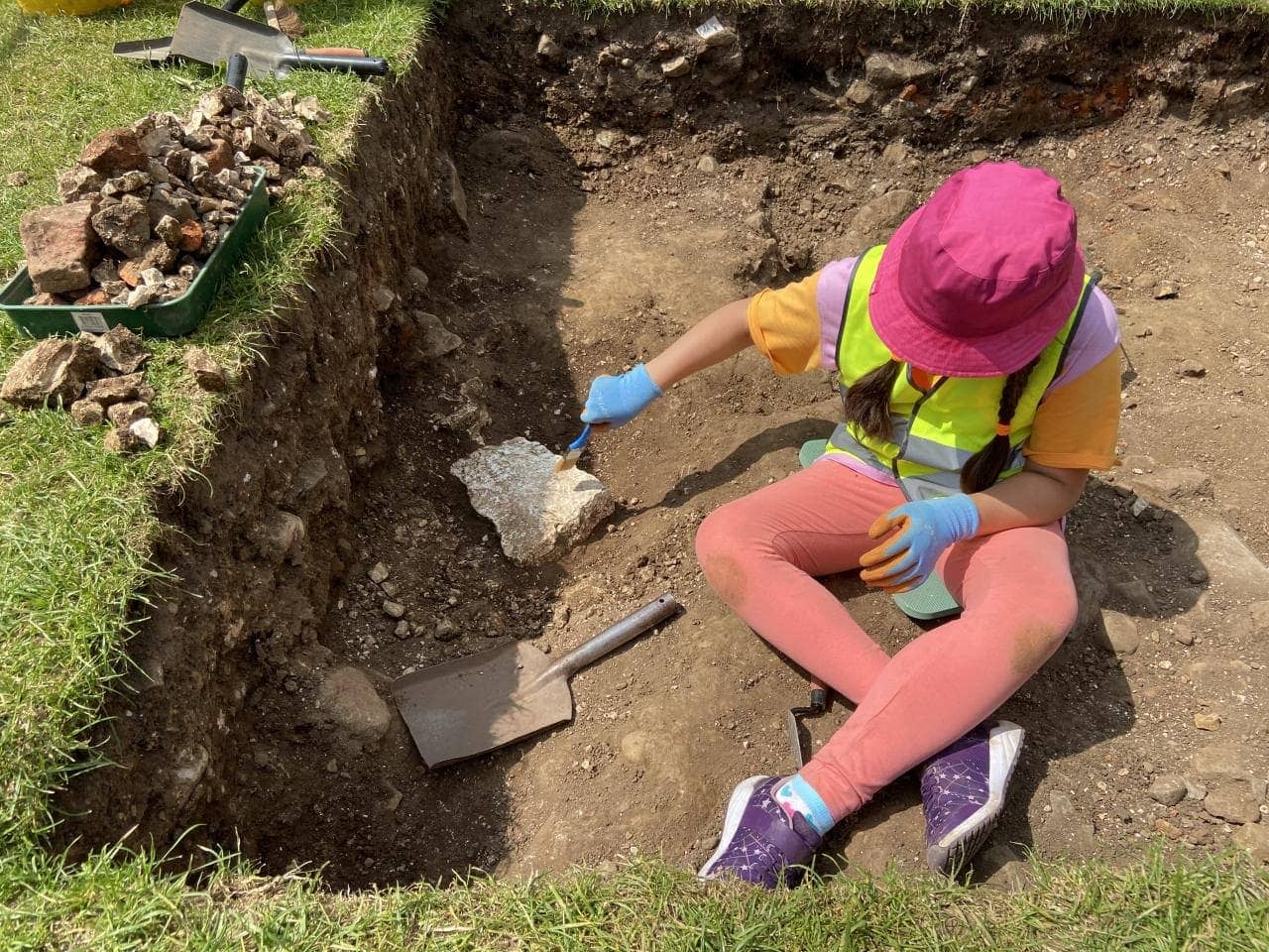 Child sits in trench trowelling area. Wears a pink bucket hat, and high vix vest