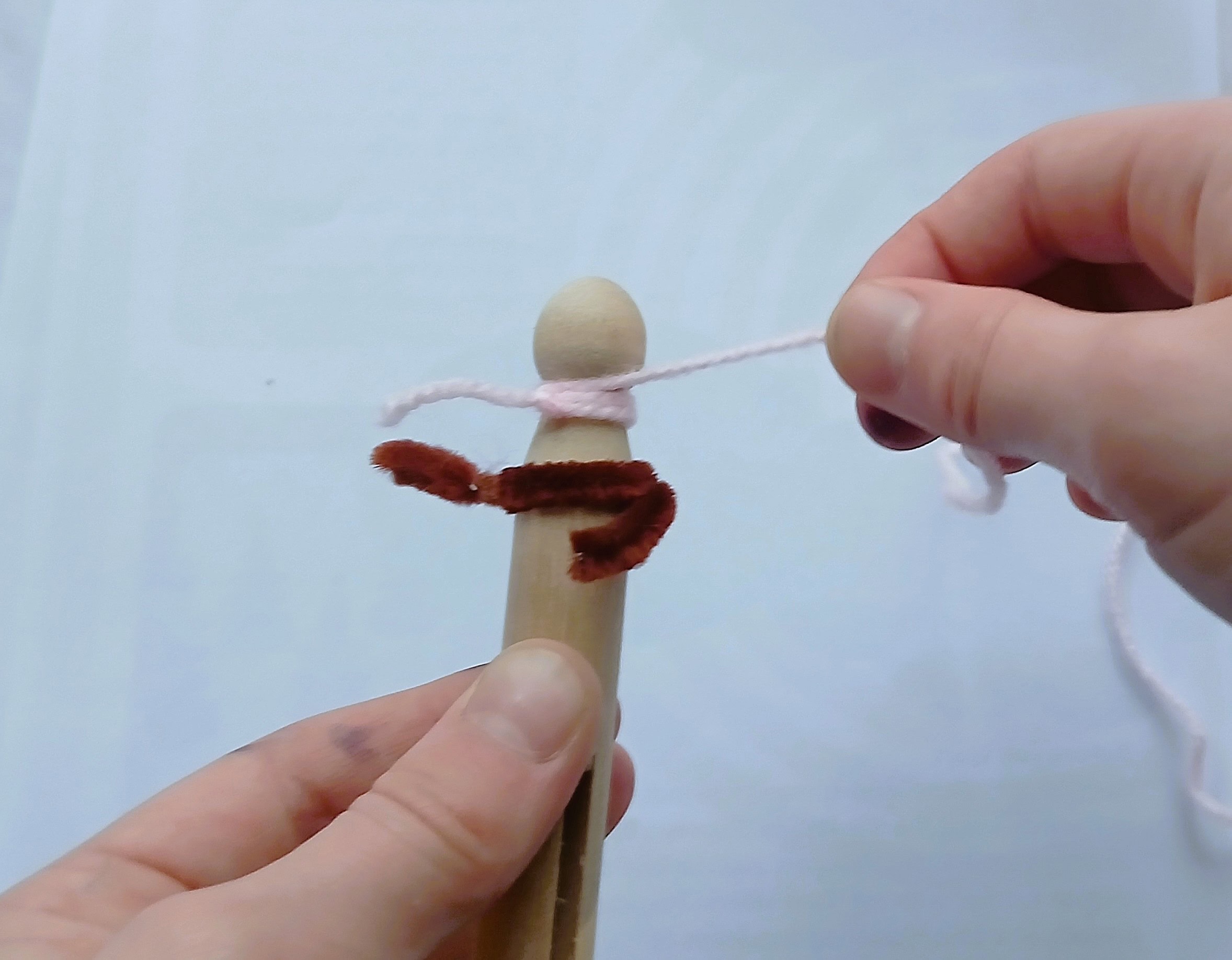 A person wraps white yarn around a wooden clothes peg with a brown pipe cleaner wrapped around it.