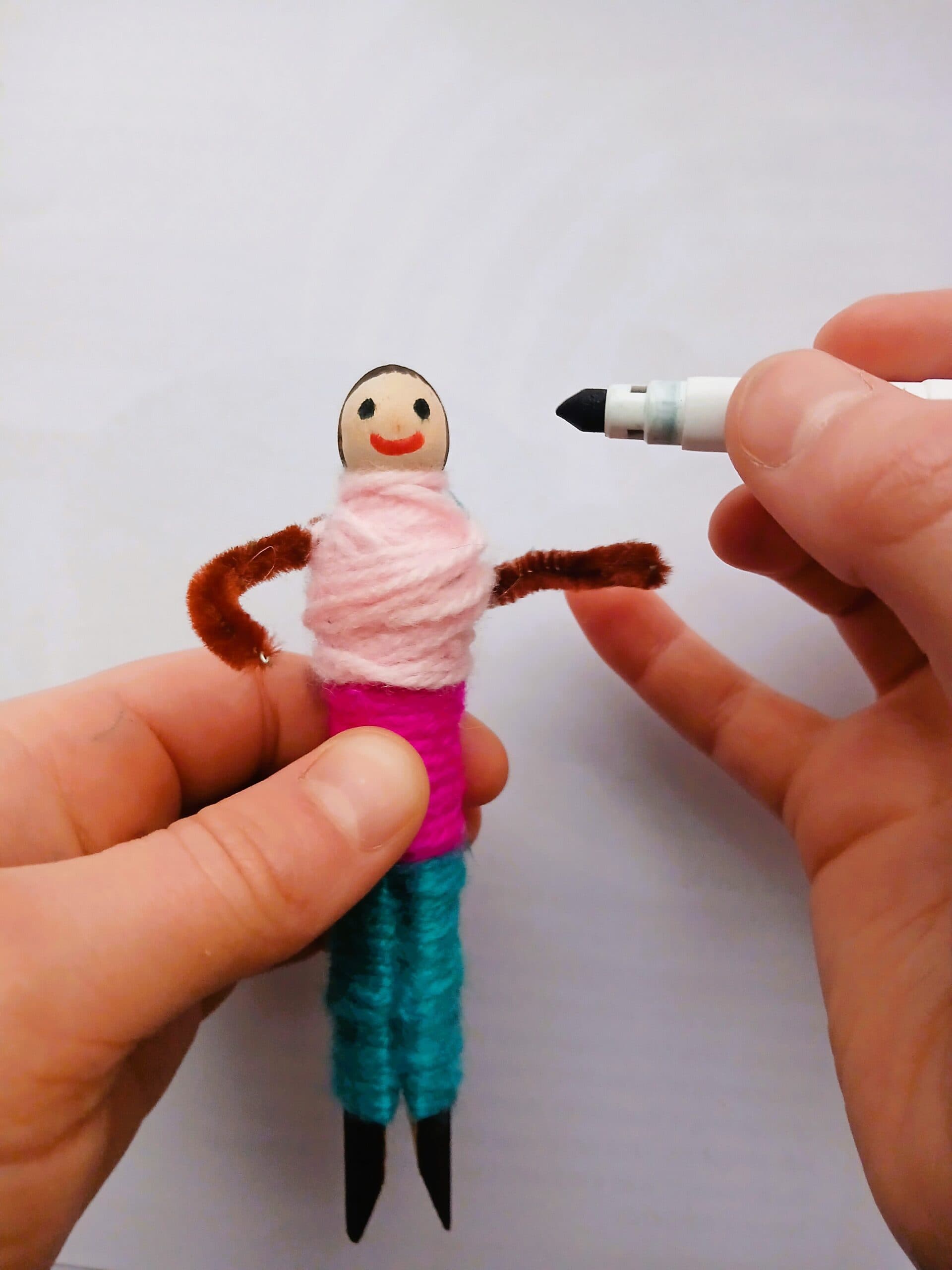 A small doll made from a peg with a smiling face, brown pipe-cleaner arms and clothes made from colourful yarn.