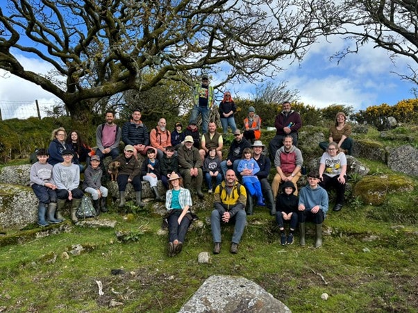 A group of 30 adults and young people sitting together on some large rocks under a tree on a sunny day.
