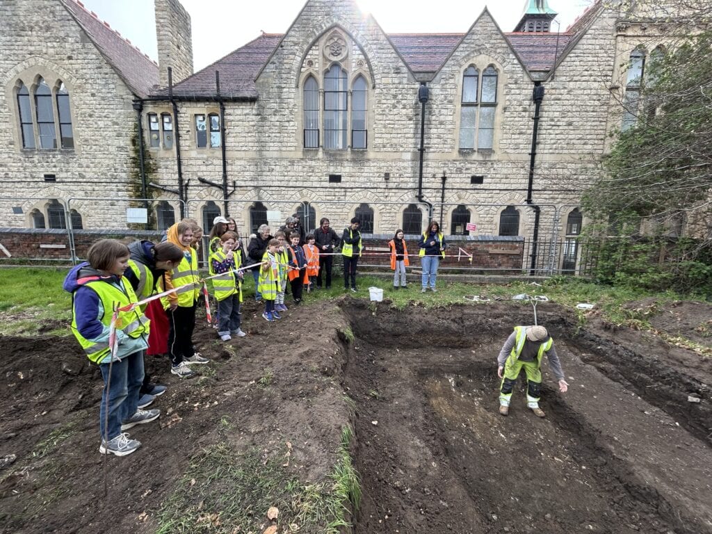 A group of children wearing high-vis vests stand around a pit on an archaeological dig. An archaeologist stands in the pit and is talking to them. It is a cloudy day and historic buildings are in the background.