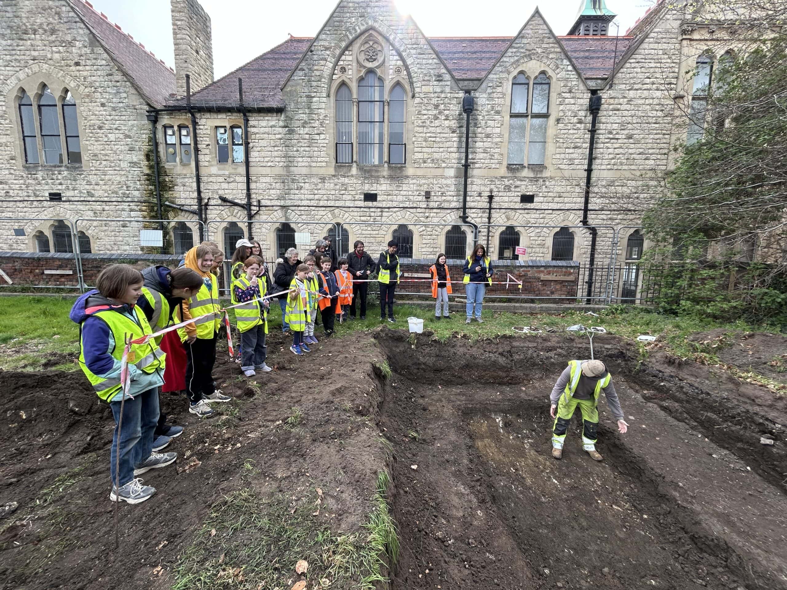 A group of children wearing high-vis vests stand around a pit on an archaeological dig. An archaeologist stands in the pit and is talking to them. It is a cloudy day and historic buildings are in the background.
