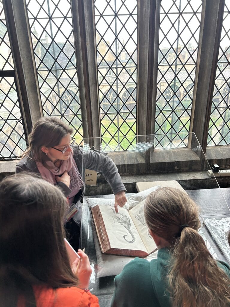 Two children sit facing an adult in a cathedral library. The adult is pointing to an open page of an old book which is protected by a plastic screen. Church windows are in the background and cathedral cloisters can be seen through the window.