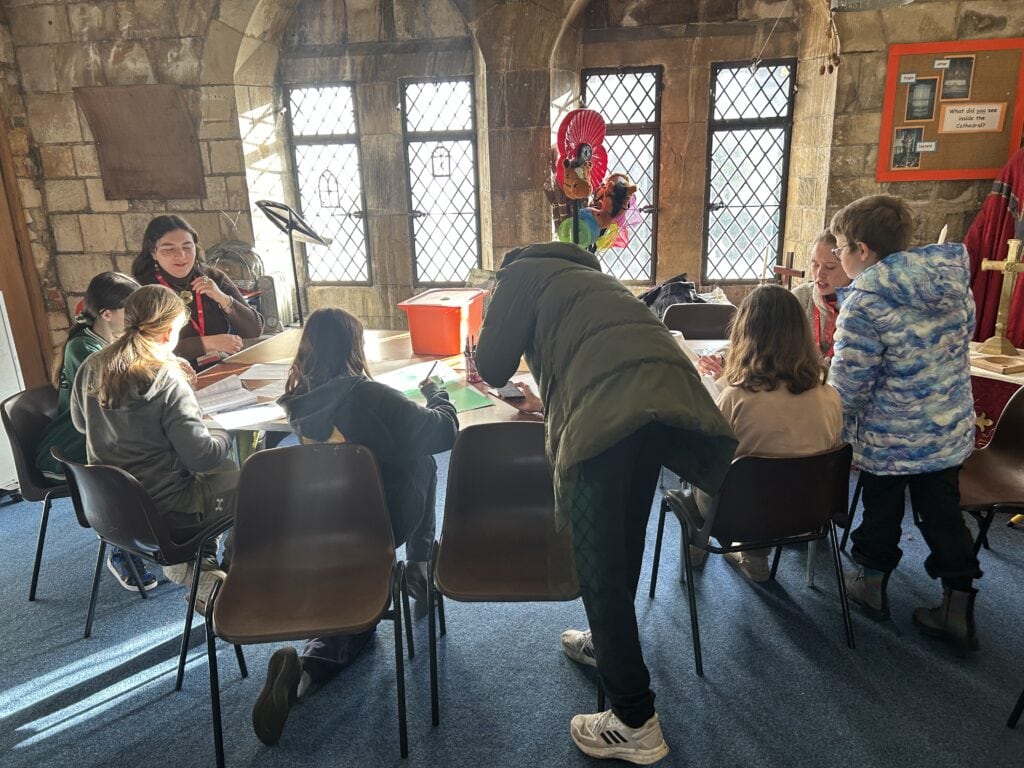 Six children and two adults sit around a table in a stone room with church windows.
