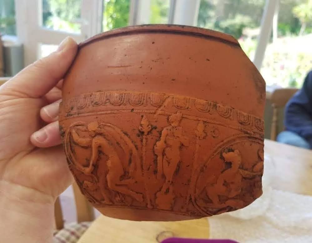 A large piece of Roman samian ware pottery. It is a shiny reddish-orange pottery fragment with Roman designs of people and plants.