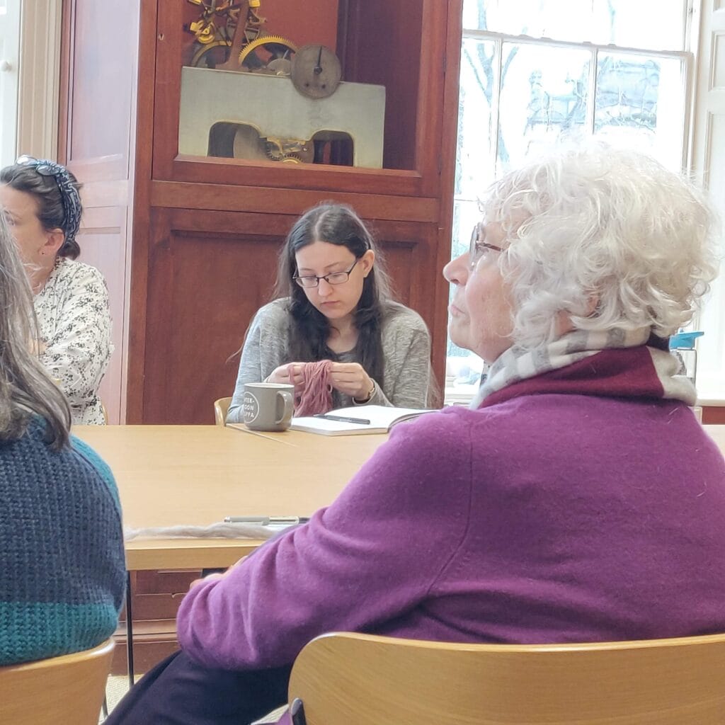 In the foreground a woman wearing a pink jumper and scarf sits at a table. Three other women are in the background, also sat around table. There is an old clock and a Georgian-style window behind them.