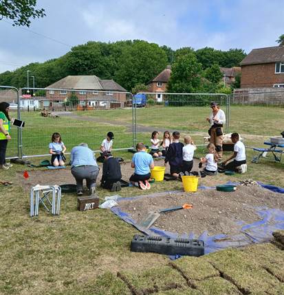 A group of children and adults working on an archaeological dig on a sunny day.