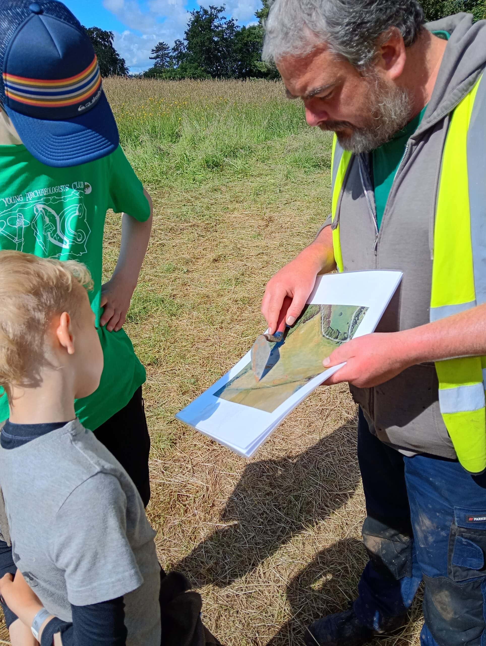 A map wearing a high-visibility vest talks to two young people standing on the edge of a field. The map is showing them an aerial photograph of some fields and pointing at the image with a metal trowel.