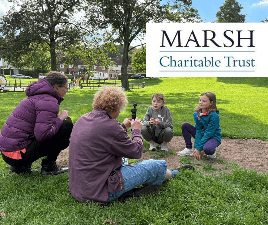 Two adults and two children sitting on the grass looking at archaeological objects. Text on a white background says: Marsh Charitable Trust.