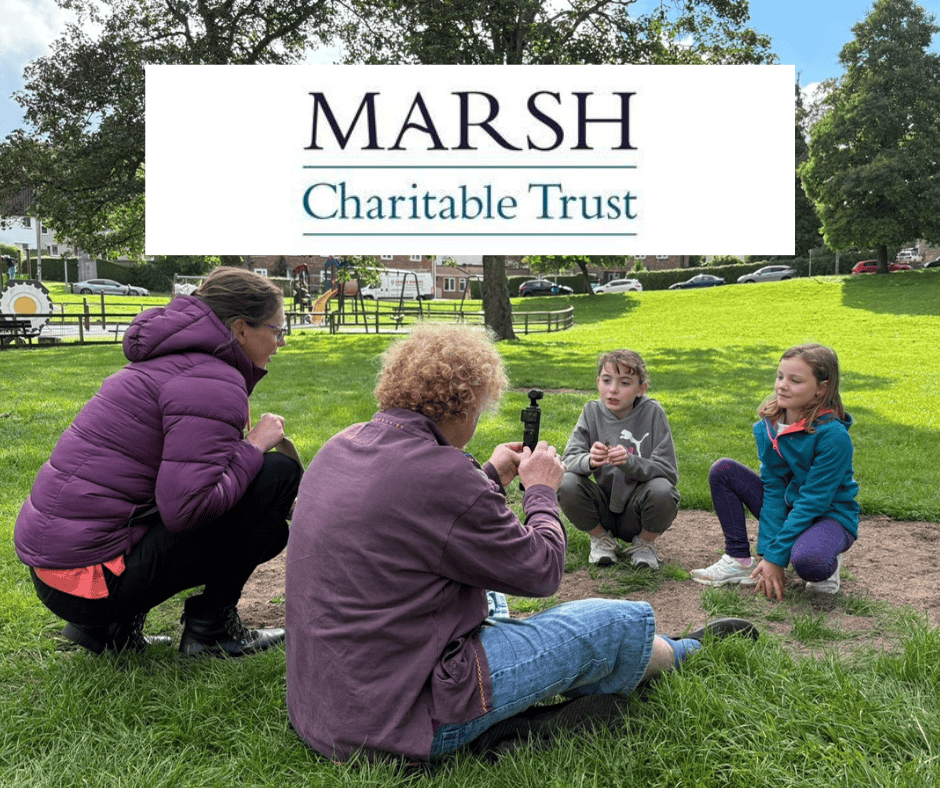 Two adults and two children sitting on the grass looking at archaeological objects. Text on a white background says: Marsh Charitable Trust.
