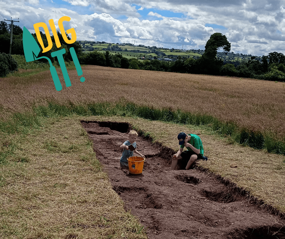 Two young people digging in a long rectangular trench on an archaeological dig. Fields and trees are in the background.