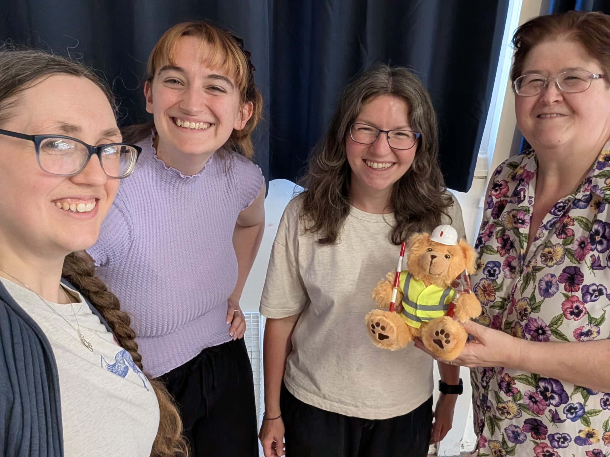 Four adult women standing in a group and smiling. One is holding a teddy bear wearing a high-vis vest and a mini hard hat.