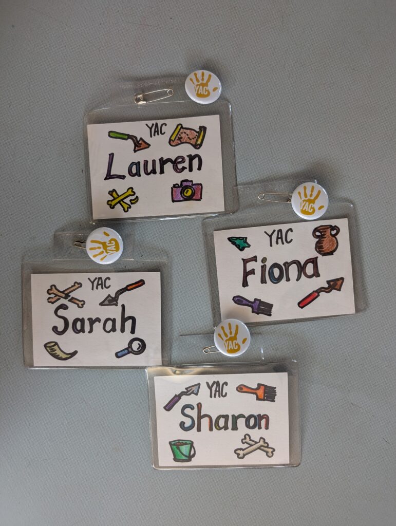 Four hand-drawn name badges reading: "Sharon, Fiona, Lauren, Sarah". They are decorated with illustrations of archaeological finds and tools.