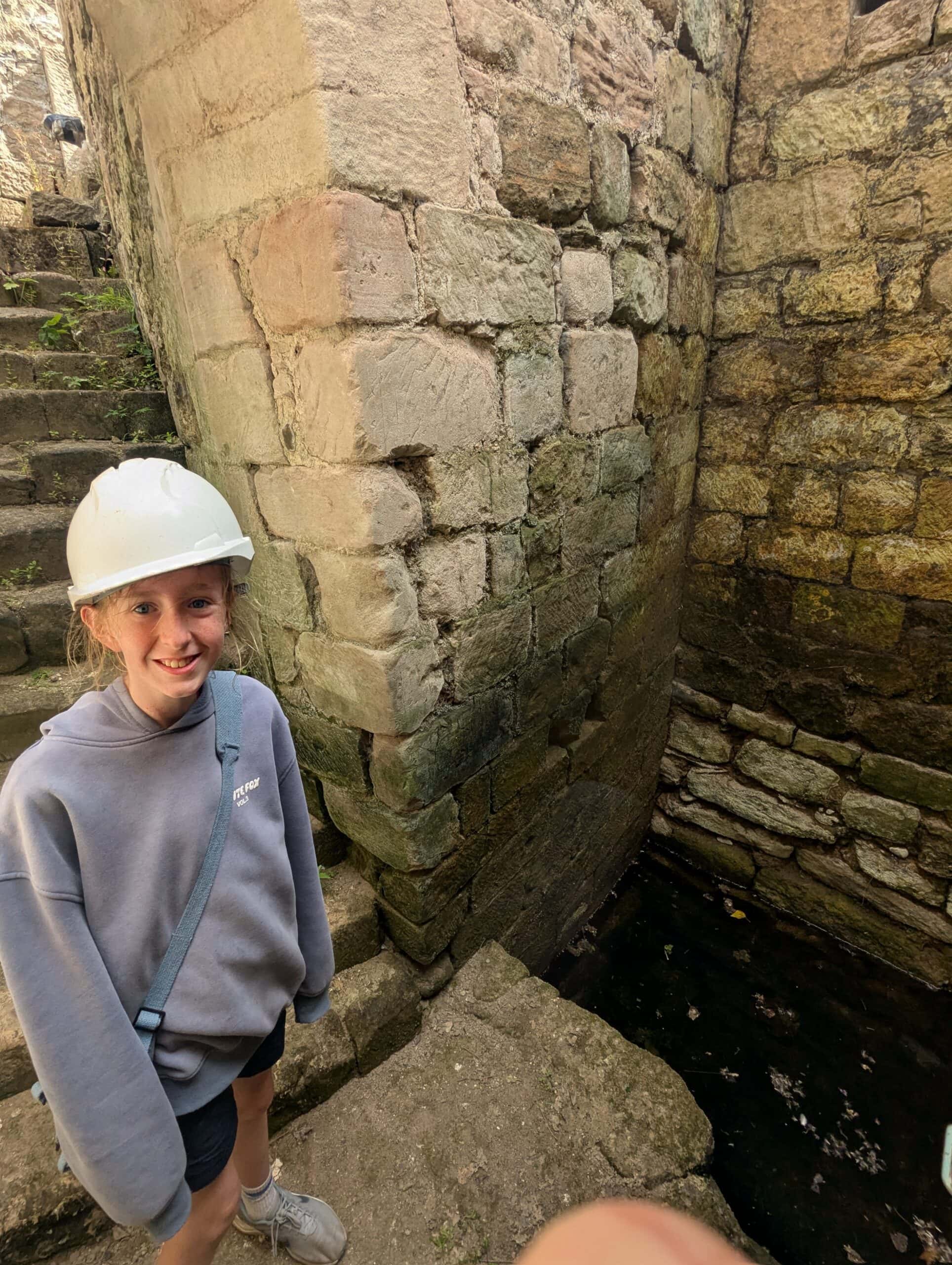 A young person wearing a white hard hat stands at the bottom of a stone staircase in the ruins of a medieval castle.