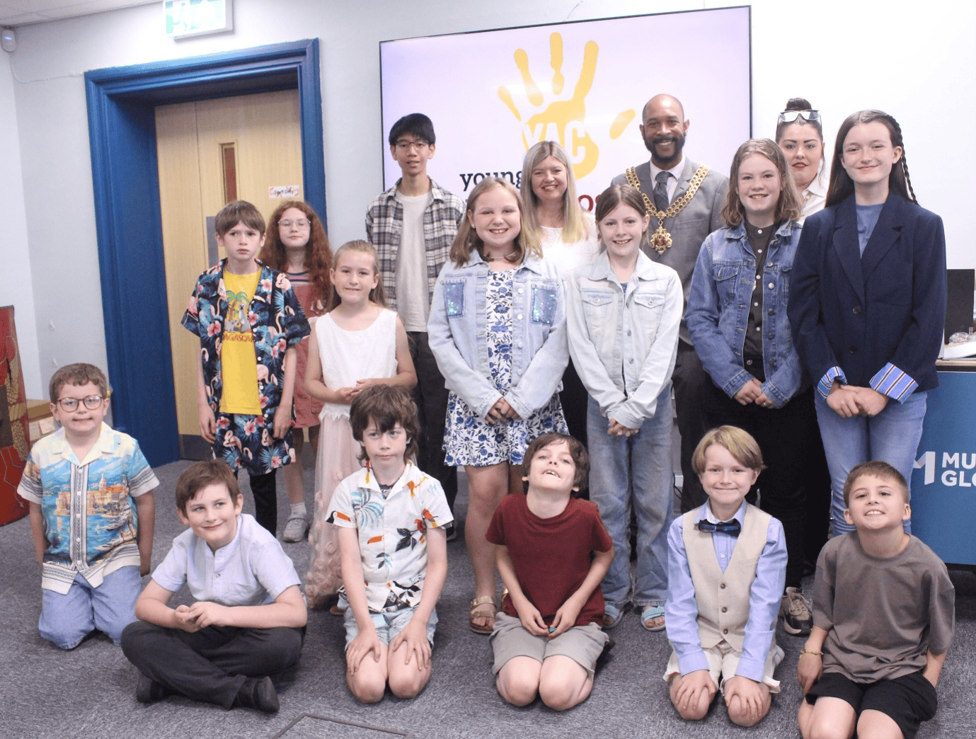16 young people aged between 8 and 16 sitting or standing in a group, accompanied by a man wearing a mayoral chain of office.