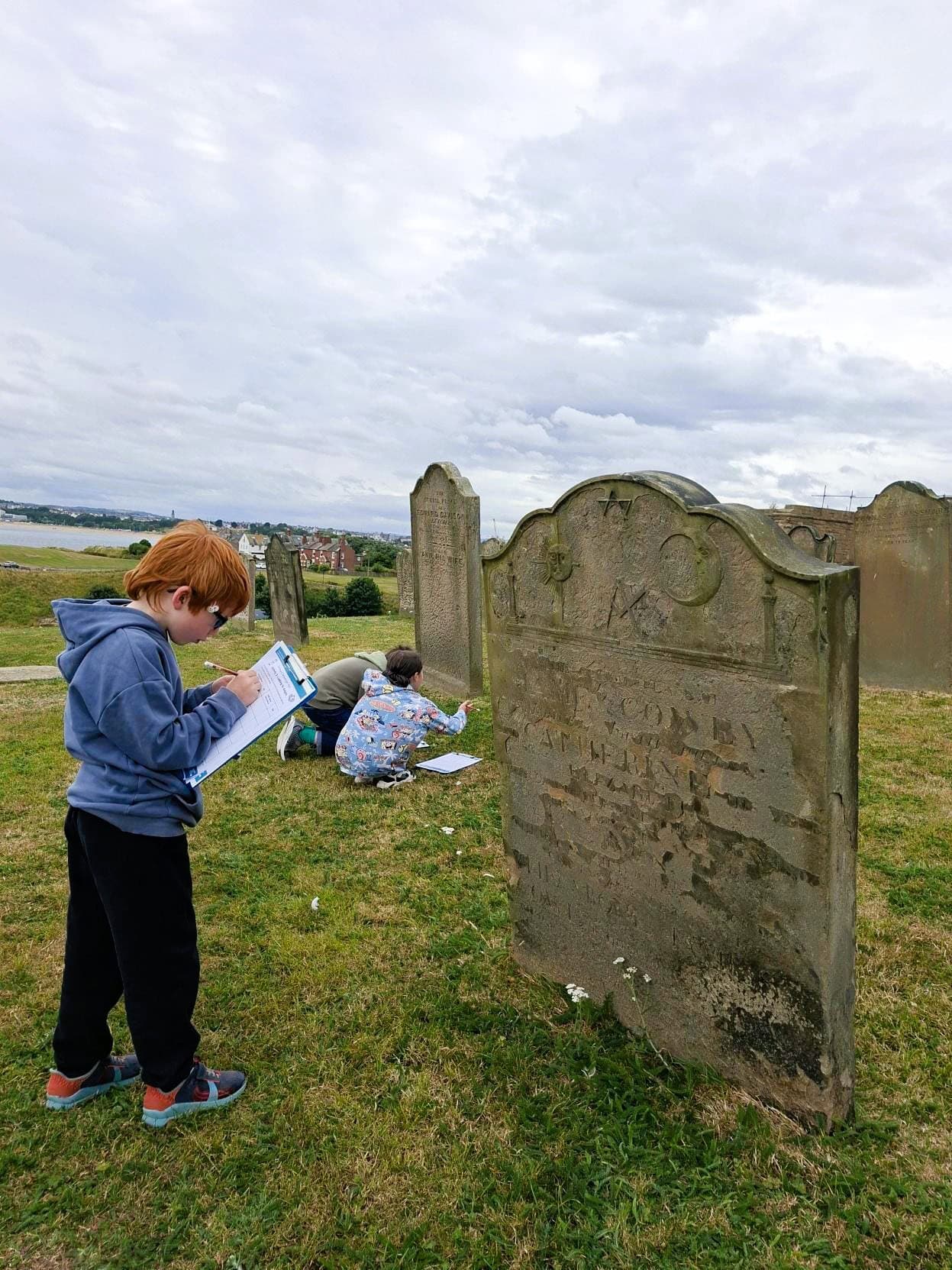 A child is standing on the grass in front of an old gravestone. He is writing on a worksheet on a clipboard with a pencil. Other gravestones are in the background.