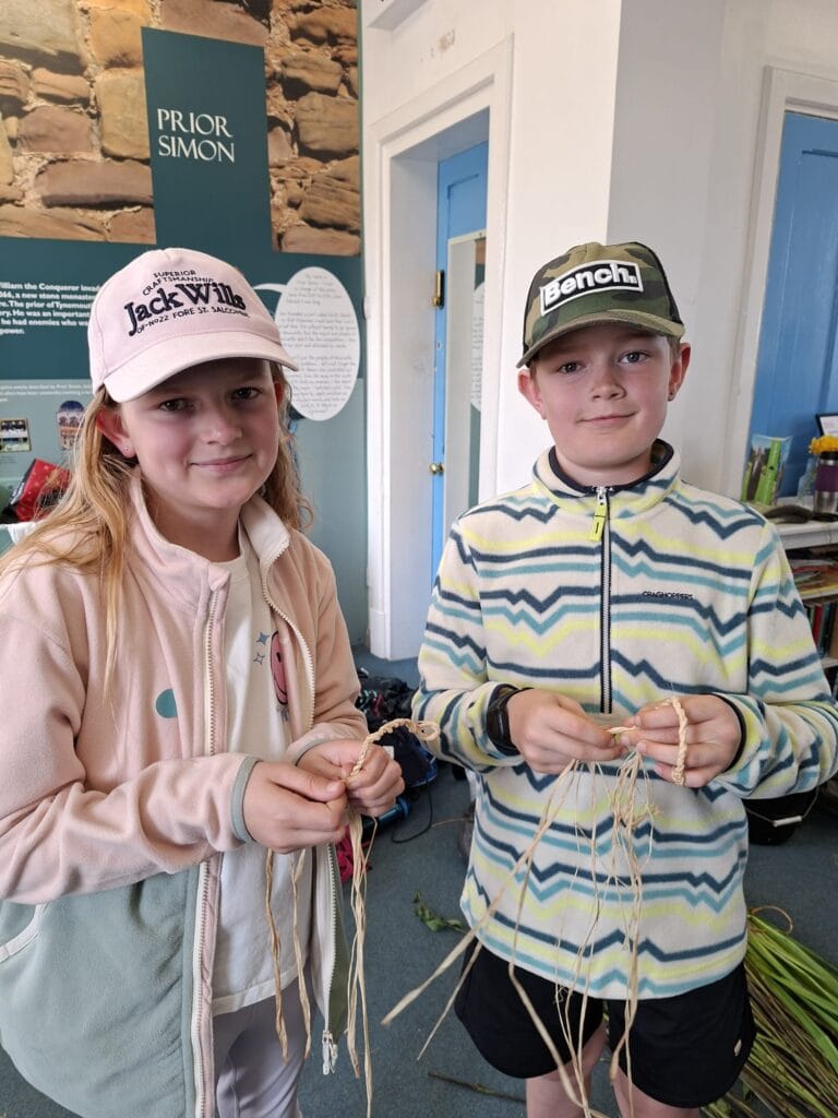 Two children wearing fleeces and caps are standing and holding cord from plant fibres.
