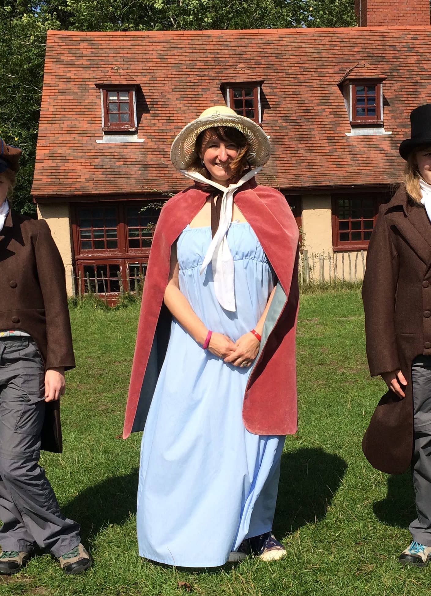 A woman standing on the grass in front of an historic house. She is wearing a bonnet and Georgian dress and cape.