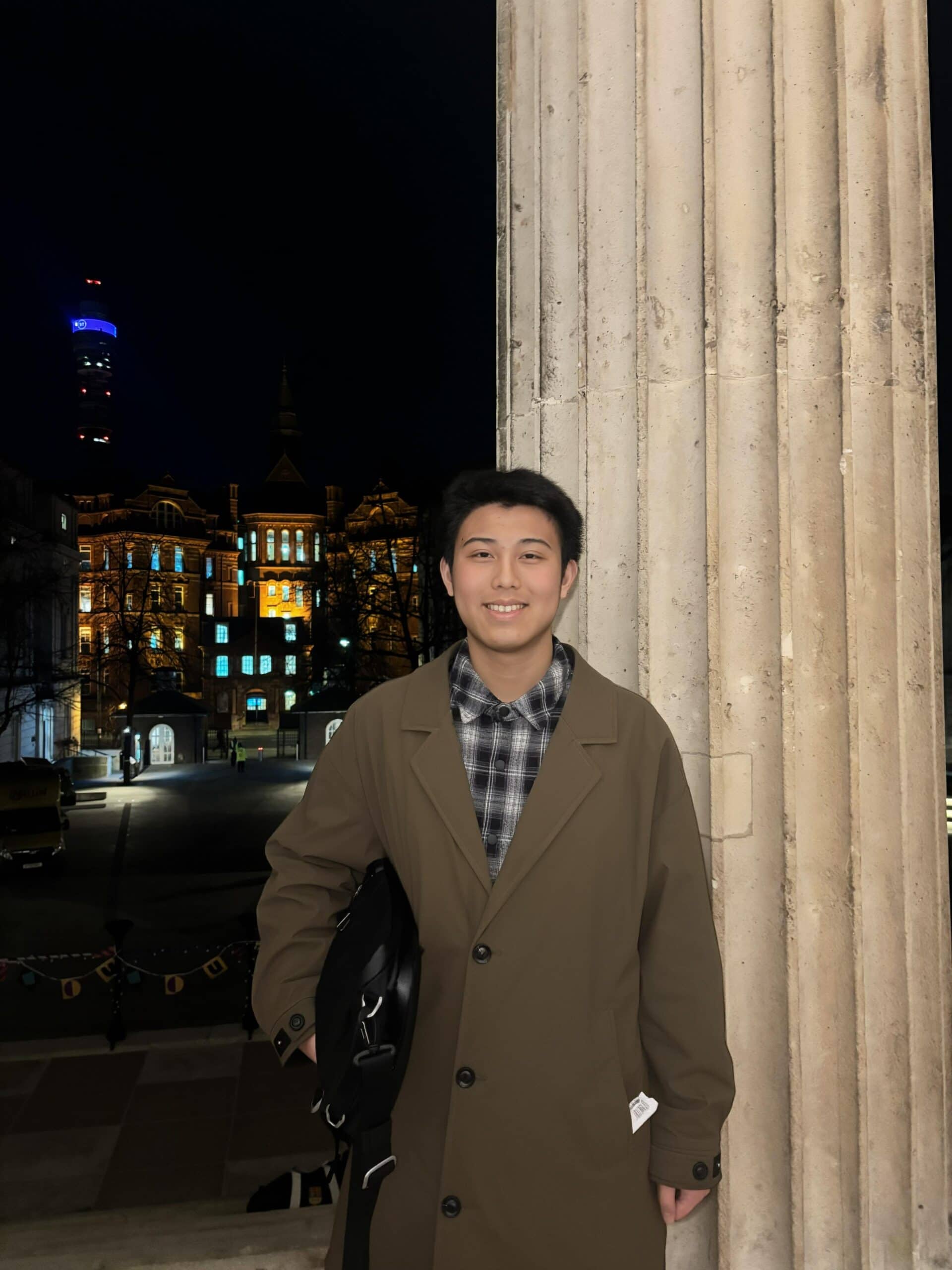 A man in his early twenties stands in front of a stone column wearing a brown coat.